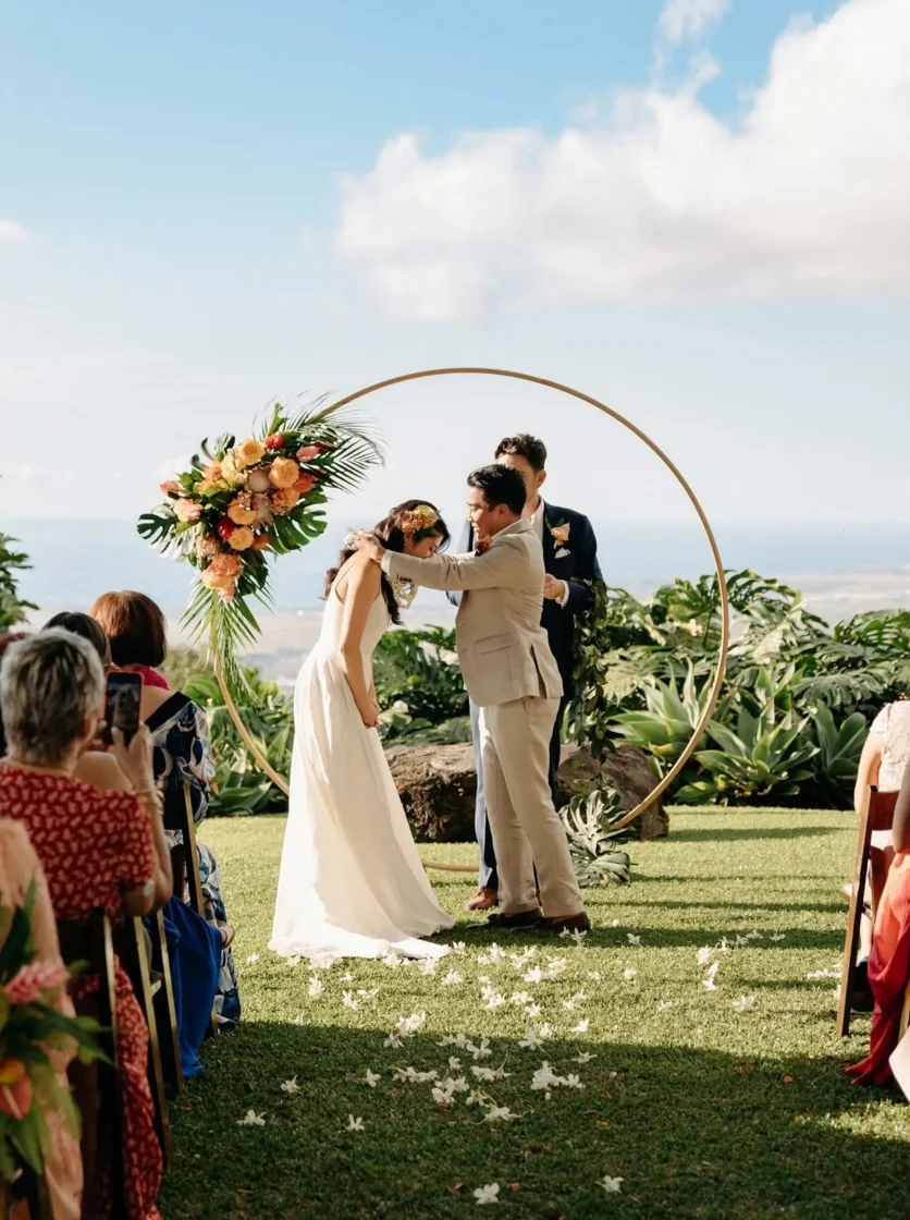 Couple exchanging vows in front of gold circle arch with ocean backdrop
