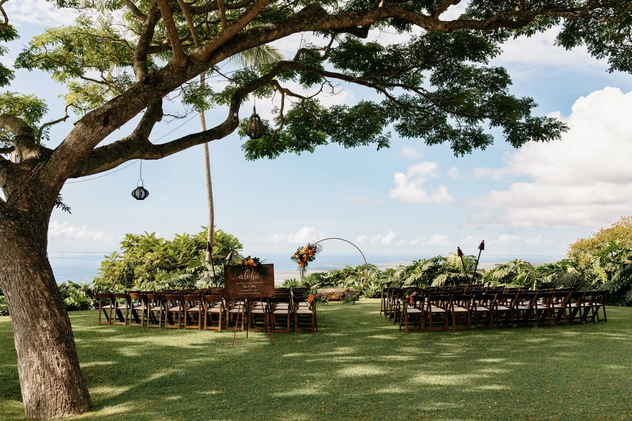 Ceremony setup under large tree with round arch and wooden chairs